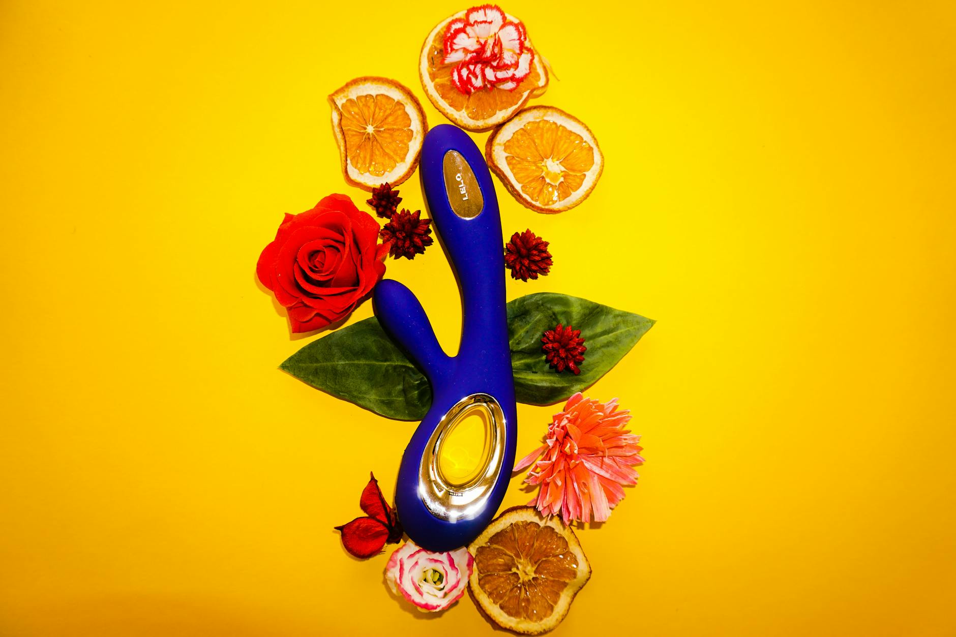 A blue clitoral vibrator surrounded by fresh flowers and citrus slices on a bright yellow background
