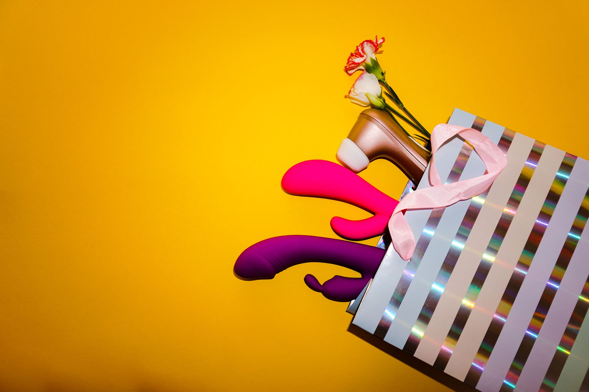 Arrangement de vibrateurs colorés avec des fleurs dans un sac cadeau holographique sur fond jaune vif